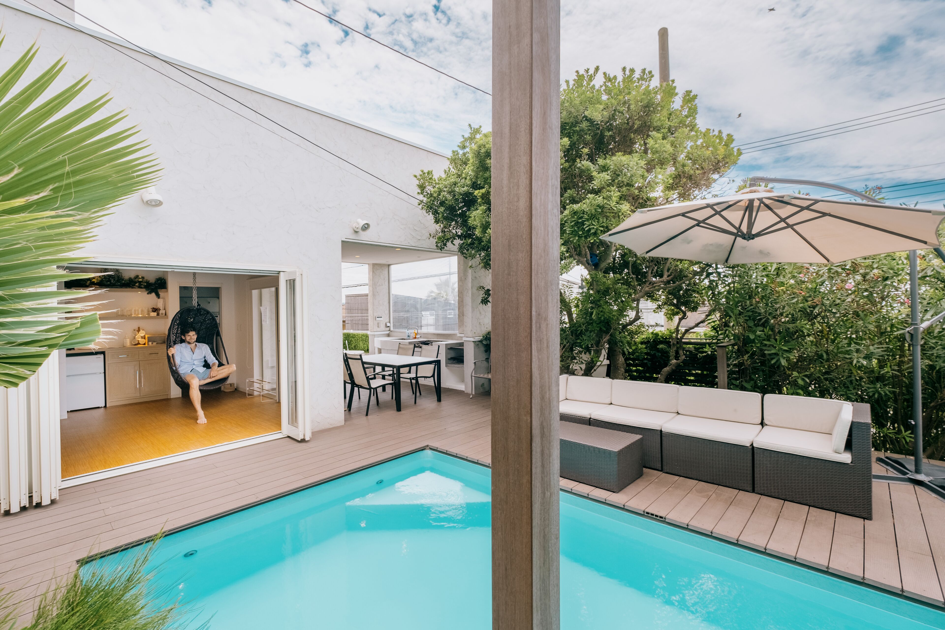 A modern outdoor pool area with a wooden deck, a seating area with a sofa and chairs, and a hanging chair.