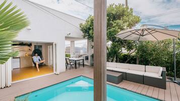 A modern outdoor pool area with a wooden deck, a seating area with a sofa and chairs, and a hanging chair.