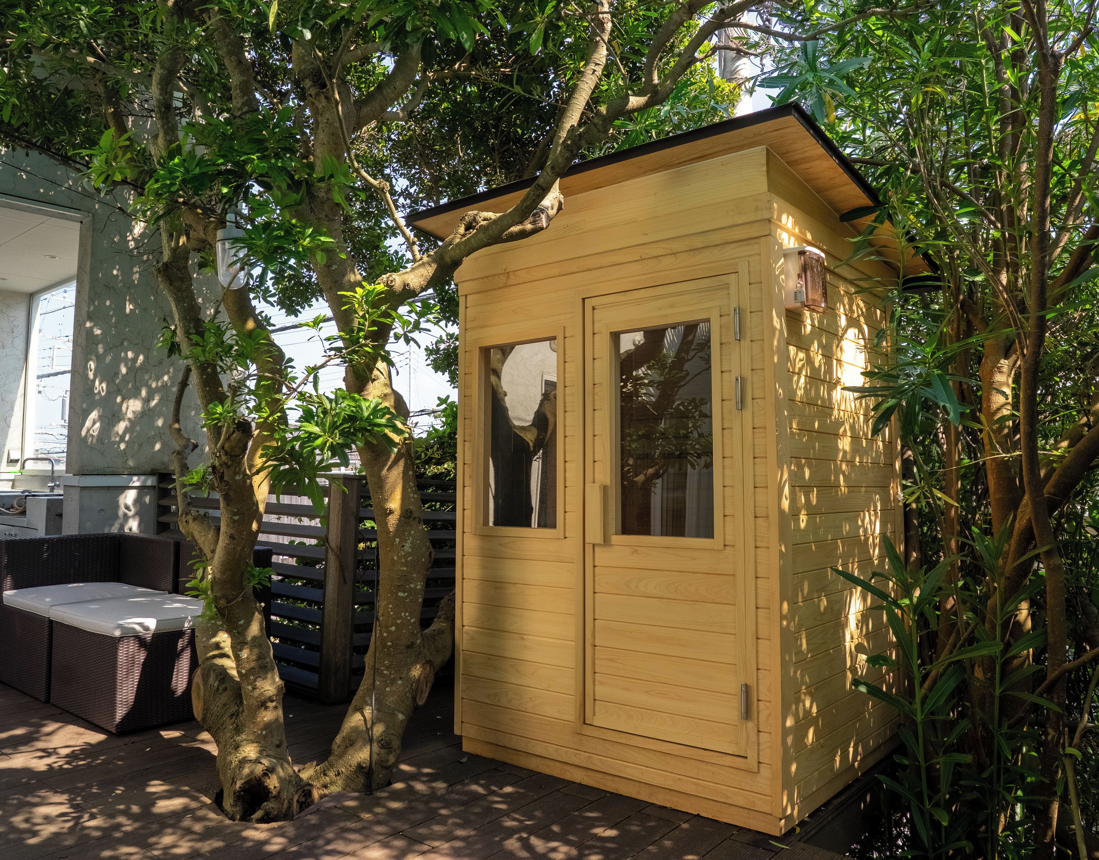 Una caseta de madera con dos ventanas, rodeada de árboles y un patio con mobiliario exterior.
