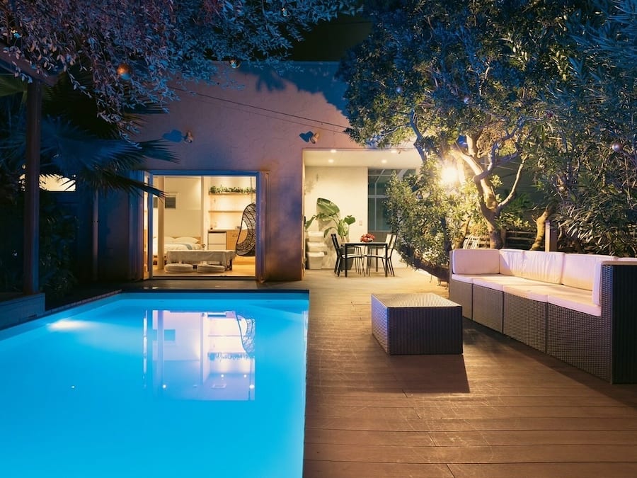 Un área de piscina con cubierta de madera, un espacio de descanso con cojines y una mesa de comedor con sillas.
