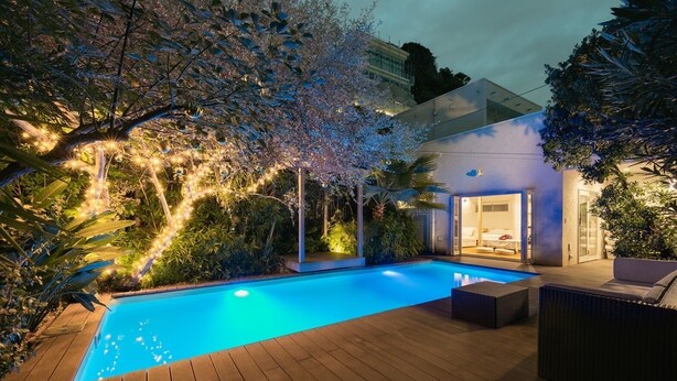 A swimming pool with a wooden deck, surrounded by lush greenery and a modern building with an open entrance. - Casablanca Pool House (Kamakura)