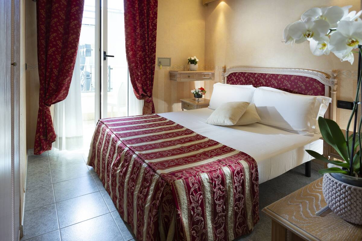 A hotel room with a large bed, a nightstand with a vase of flowers, and a potted plant on a side table.