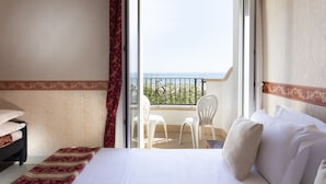 A hotel room with a bed, a nightstand, a window with a view of the sea, and a balcony with chairs.