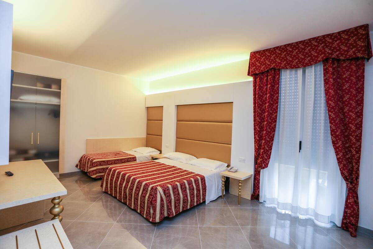 A hotel room with two beds, a desk, a wardrobe, and a large window with curtains.