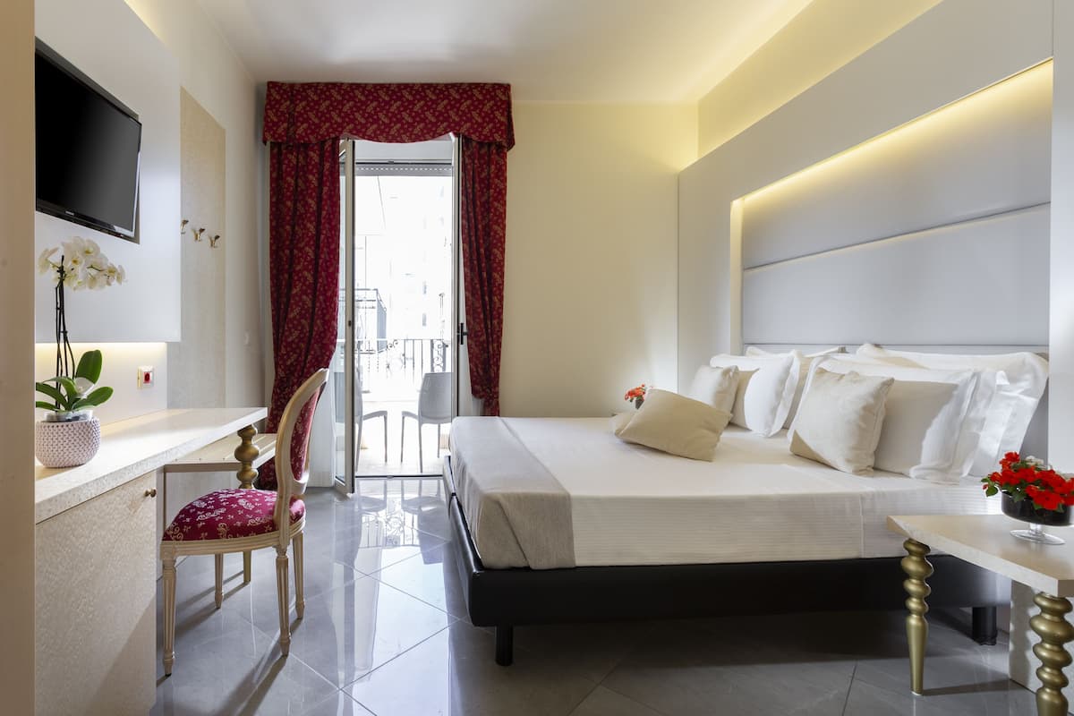 A modern hotel room with a bed, a desk, a chair, a TV, and a balcony with red curtains.