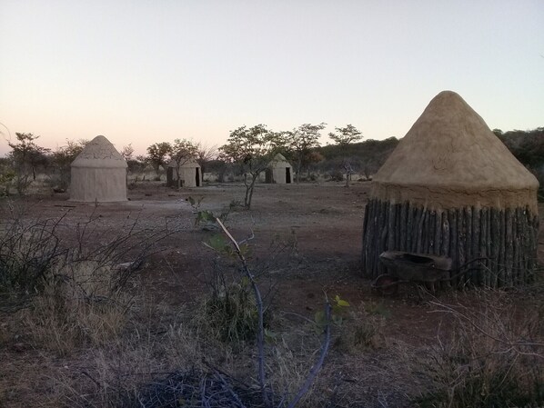 Paisaje rural con varias pequeñas chozas cónicas hechas de barro o arcilla, rodeado de vegetación escasa y árboles.