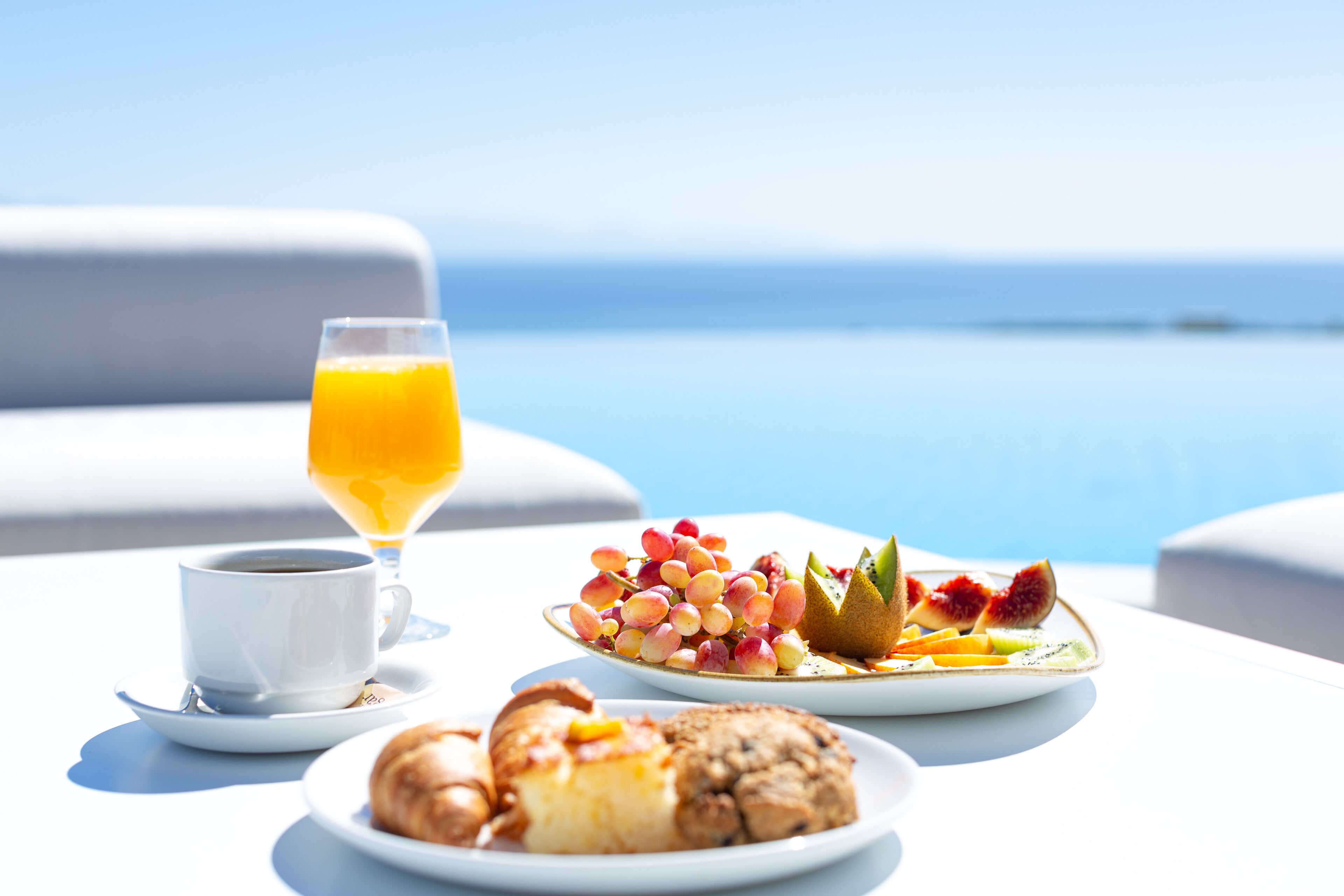 Un desayuno con una taza de café, un vaso de jugo de naranja y platos de pasteles y fruta sobre una mesa con vista al océano.