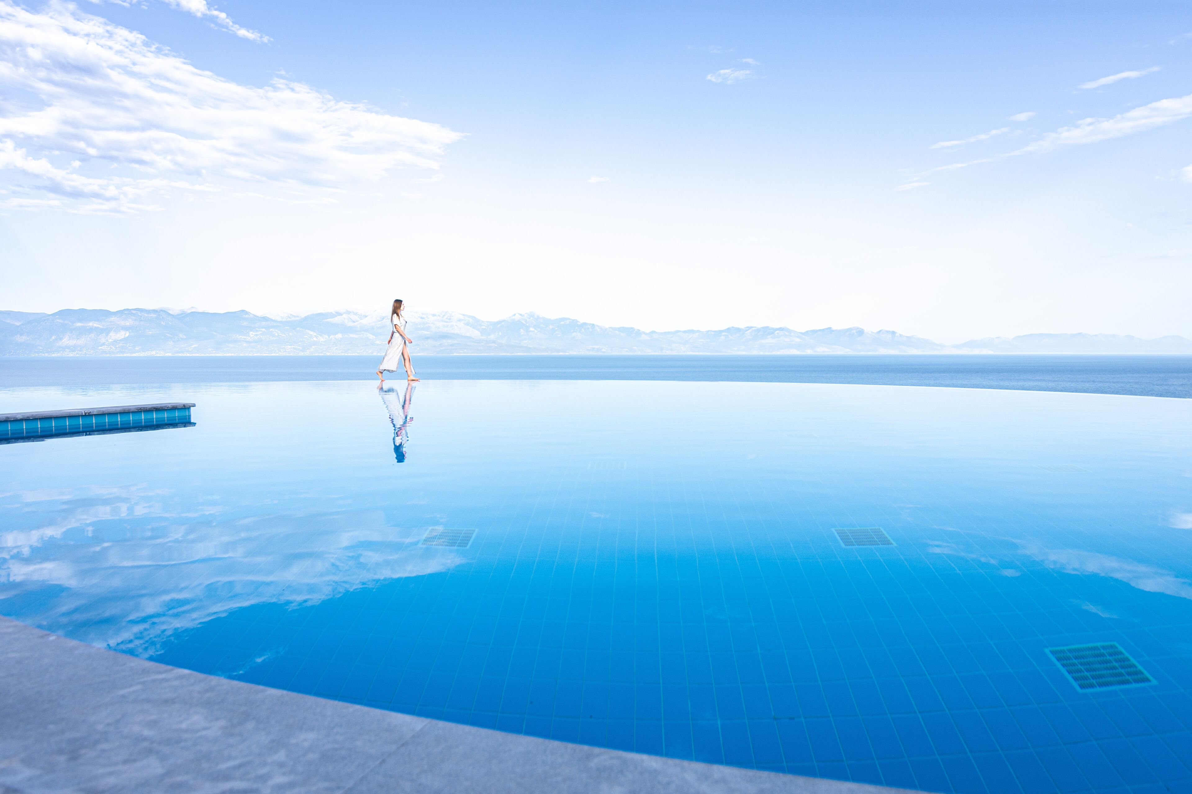 A person standing on the edge of a large, clear blue infinity pool with a mountainous backdrop.