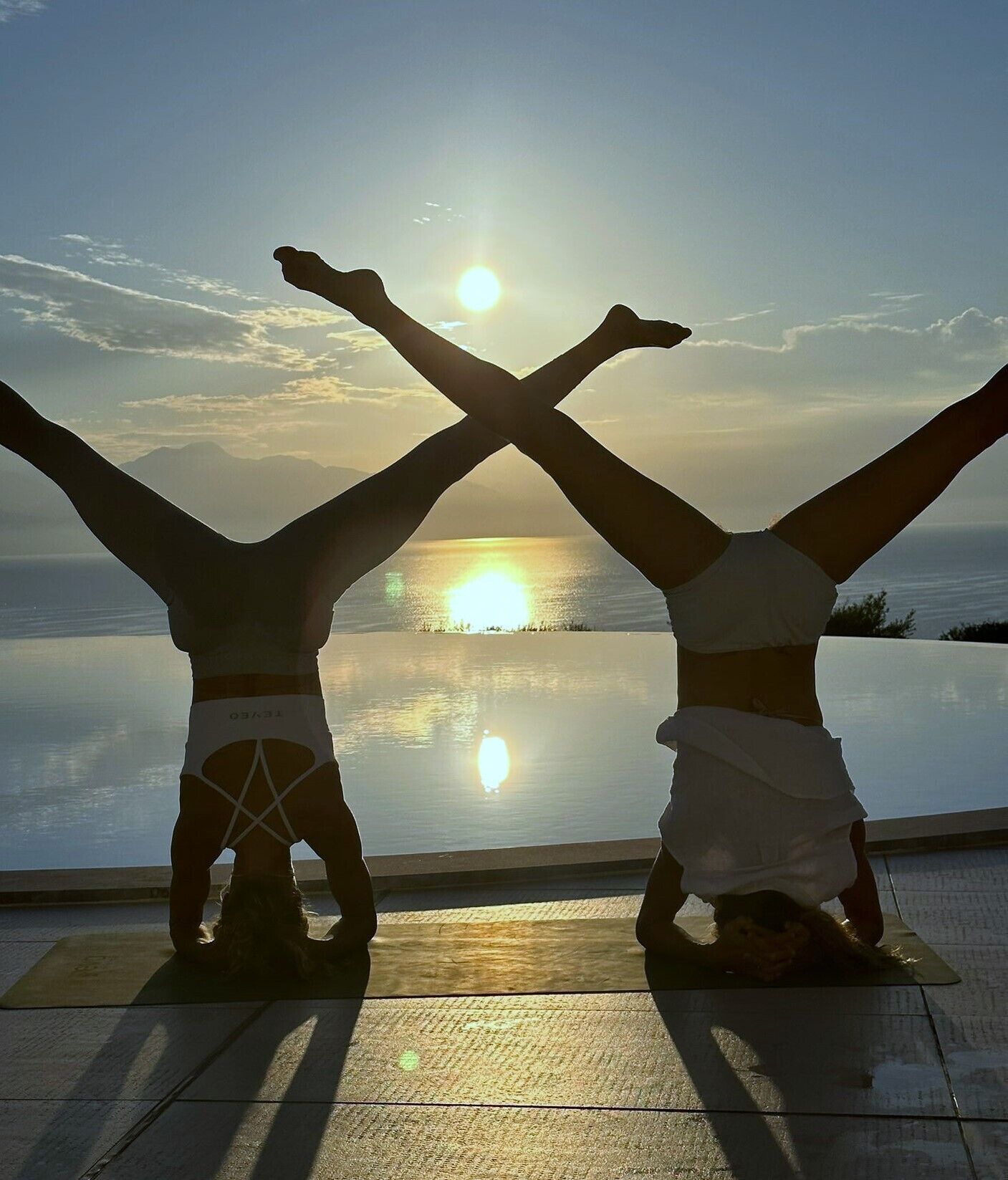 Zwei Personen führen eine Handstand-Yoga-Pose auf einer Yogamatte aus, im Hintergrund untergehende Sonne und ruhiges Meer.