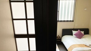 A bedroom with a bed, a nightstand, a window with blinds, and a view of a wooden staircase. - Hotel Zipangu Murasakino (Kyoto)