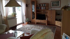 A living room with a sofa, a coffee table, a bookshelf, a television, and a window with curtains. - Max Kuehlungsborn House (Kühlungsborn)