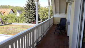 A covered balcony with a white railing, a black chair, and a view of a green roof garden and trees. - Max Kuehlungsborn House (Kühlungsborn)