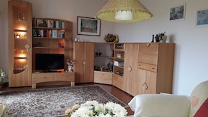 A living room with a TV, wooden cabinets, a patterned rug, and a white chair. - Max Kuehlungsborn House (Kühlungsborn)