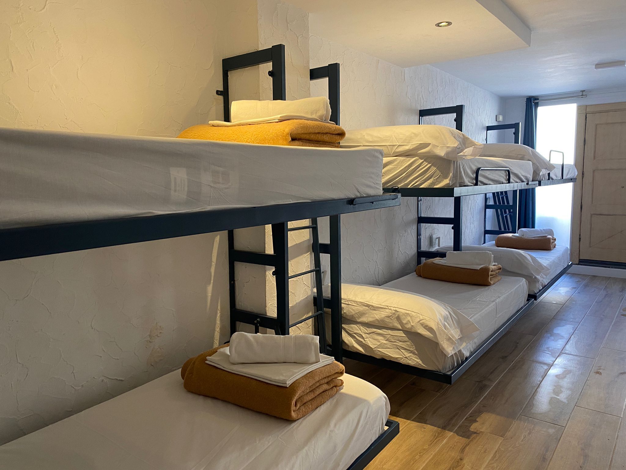 A dormitory-style room with bunk beds, wooden flooring, and a door leading to another room.