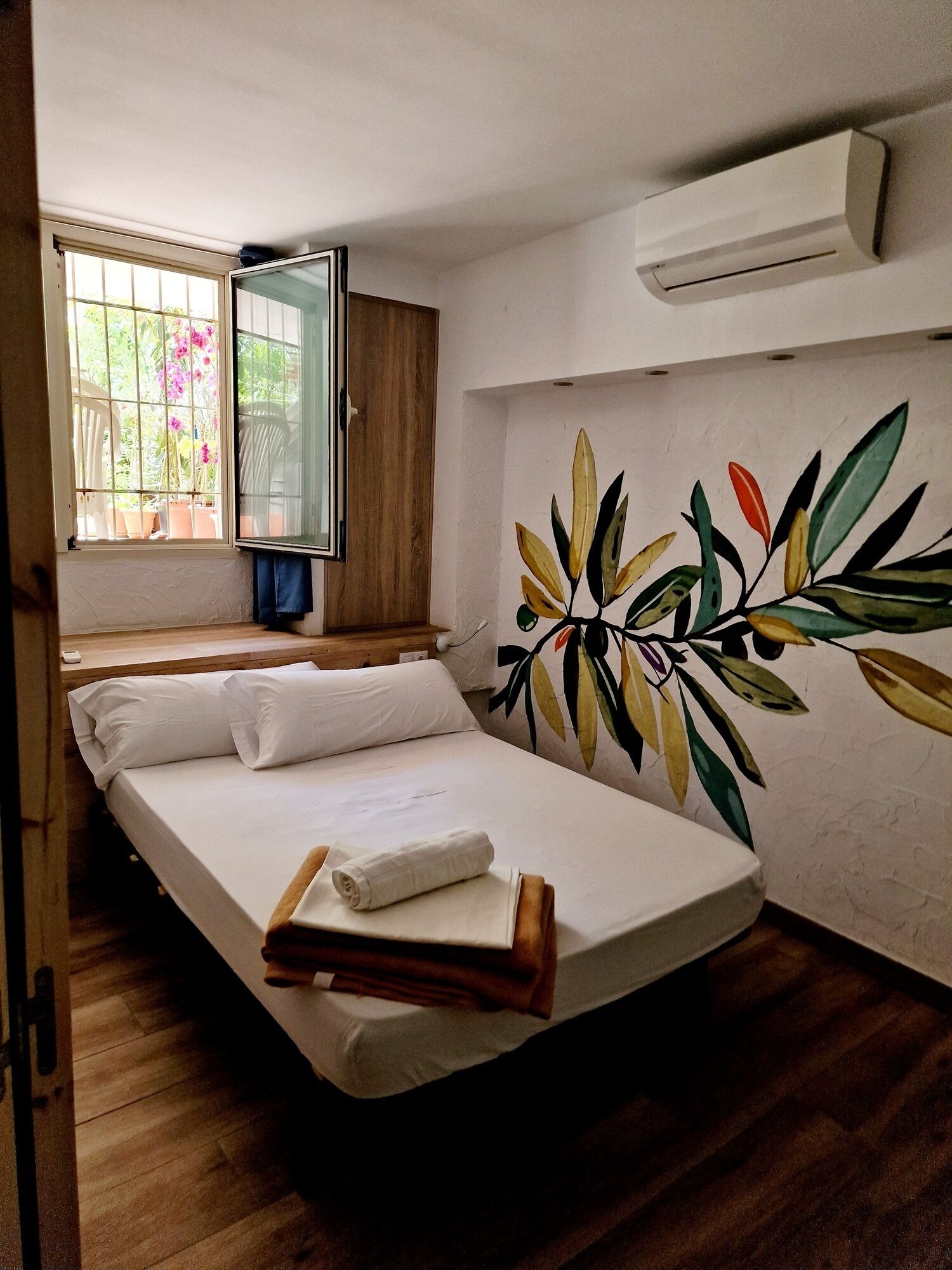 Un lit simple avec du linge de lit blanc, une fenêtre donnant sur un paysage verdoyant et un mur orné d’un motif de feuilles.
