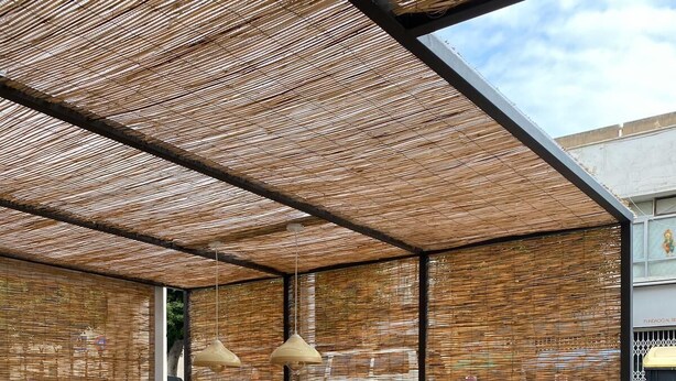 Un espacio al aire libre con mesa y bancos de madera, un toldo tejido y vistas a la calle.
