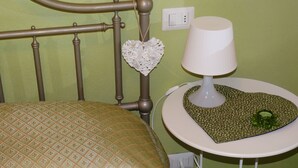 A bed with a patterned headboard, a bedside table with a heart-shaped mat and a lamp, and a power outlet on the wall.