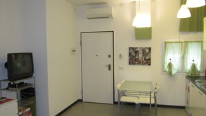 A small, sparsely furnished room with a television on a stand, a door, a table with chairs, and a window with curtains.