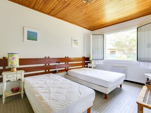 A bedroom with two beds, wooden headboard, bedside tables, a lamp, and a window with a view of trees.