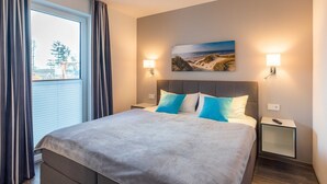 A hotel room with a large bed, two bedside tables, a nightstand, a painting on the wall, and a window with curtains. - Exclusive apartment with sea view near Kaiserbädern - Windgott (6-1) (Garz/Usedom)