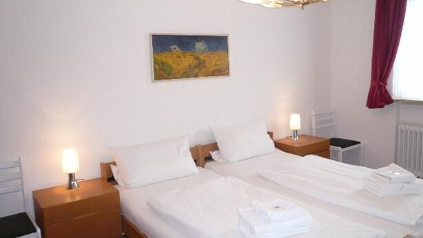 A double bed with white linens, two bedside tables with lamps, a painting on the wall, and a window with a red curtain.