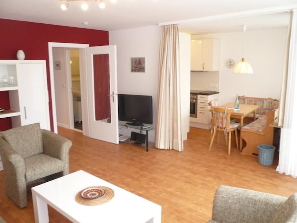 Una sala de estar con televisor, un comedor con mesa y sillas, y una cocina al fondo.