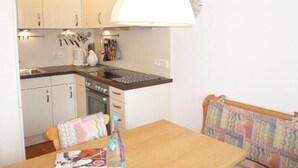 A compact kitchen with white cabinets, a stove, and a sink. There's a dining table with chairs and a colorful cushion.