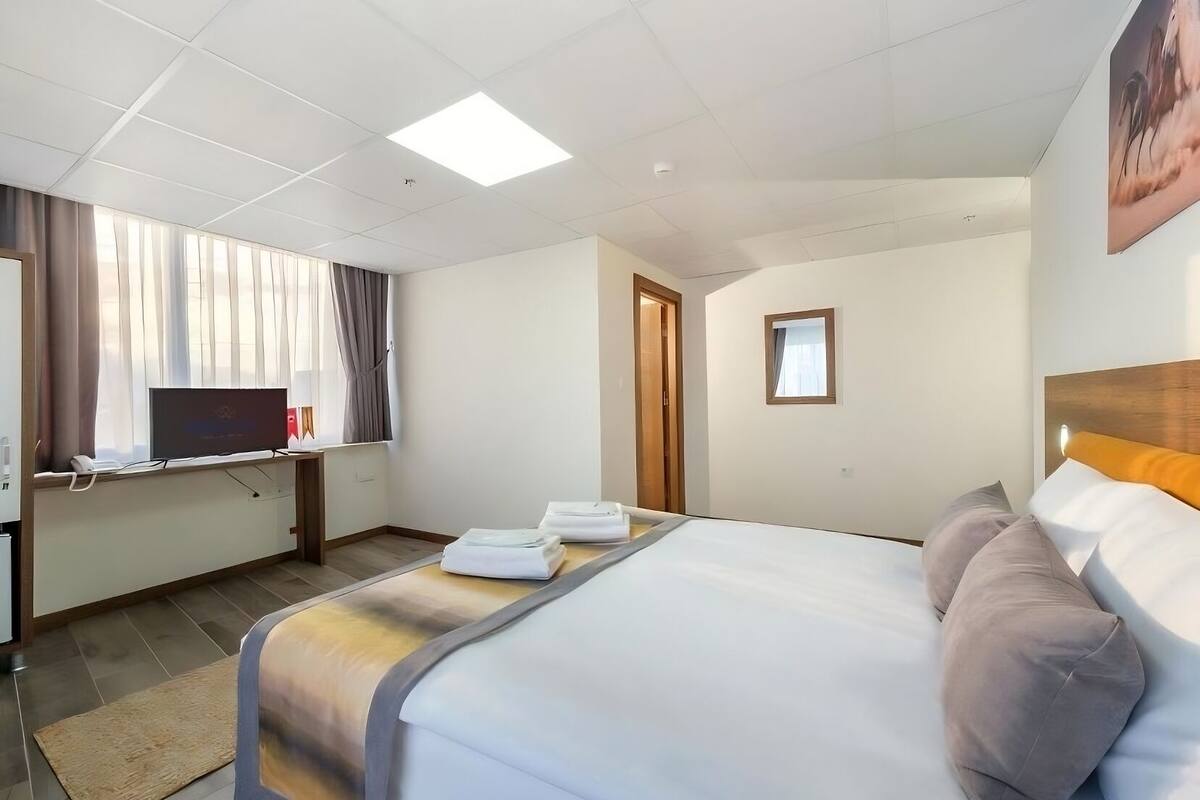 Habitación de hotel con cama, escritorio, computadora, televisor y ventana con cortinas.