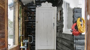 Eine Holzhütte mit weißer Tür, einem kleinen Teppich, einem Fahrrad und einem rot-schwarzen Kissen auf einem Hocker.