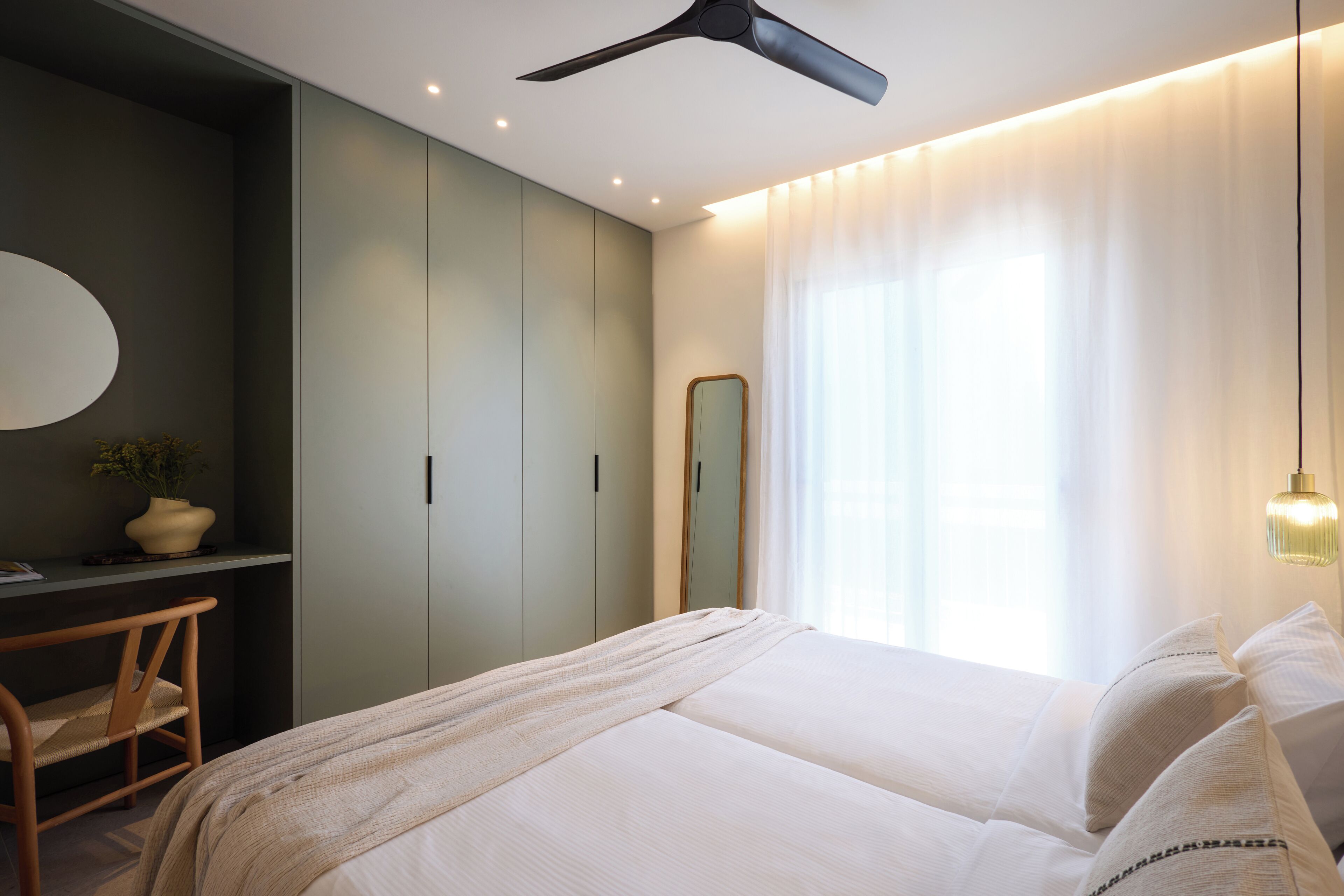 A bedroom with a bed, a desk, a chair, a mirror, and a ceiling fan.