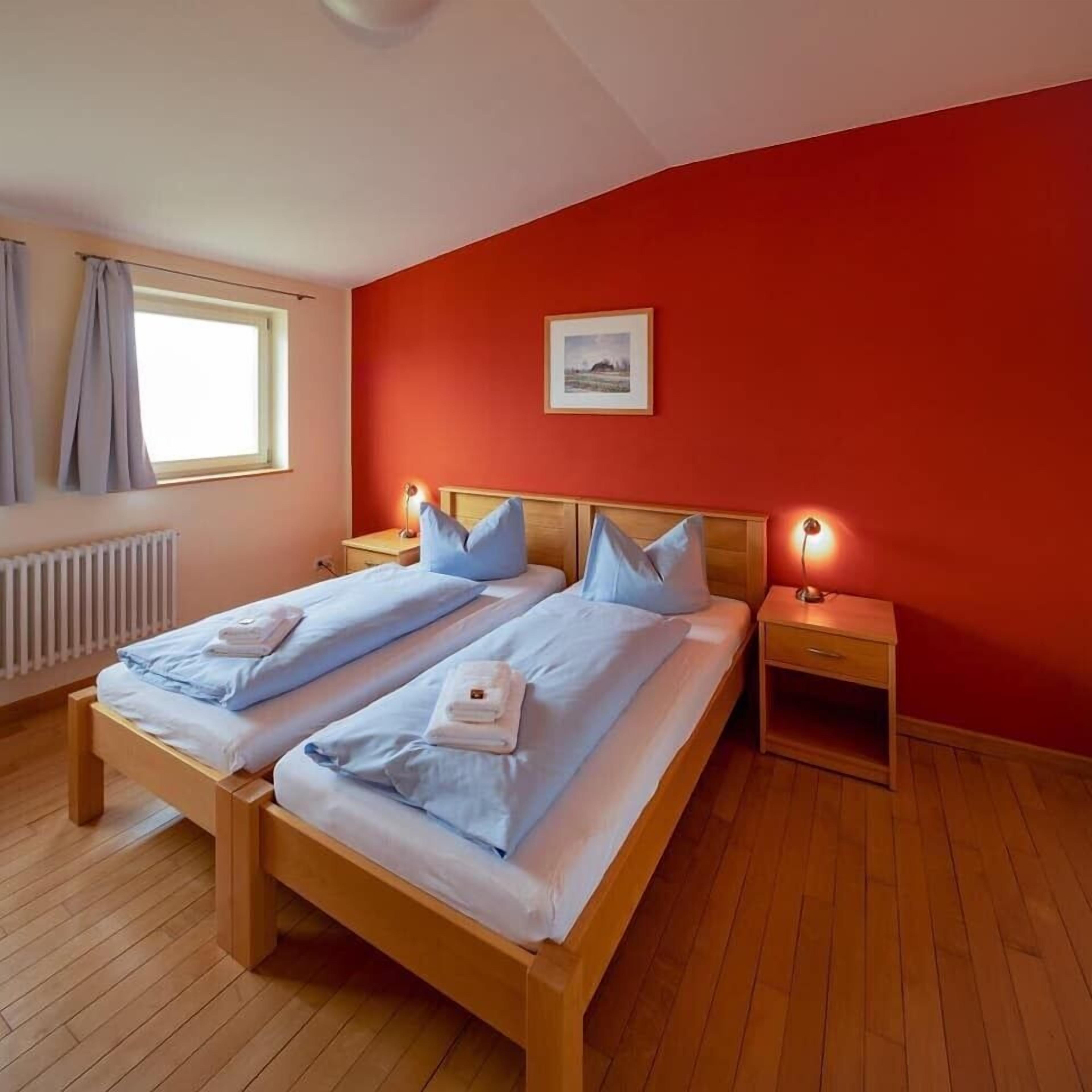 A double bed with blue bedding, two bedside tables with lamps, a window with curtains, and a radiator.