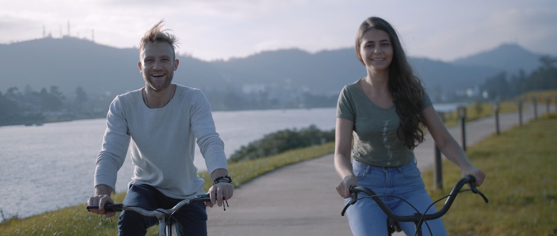 Two people riding bicycles on a path beside a body of water with mountains in the background.