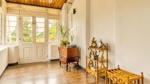 A room with wooden flooring, a wooden ceiling, a white wall, a wooden cabinet, a wooden side table with a clock, and a window with a view of trees. - The Clovelly Bungalow (Nuwara Eliya)