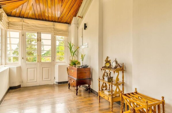 Una habitación con suelo y techo de madera, pared blanca, un armario de madera, una mesita de noche con un reloj y una ventana con vistas a árboles.