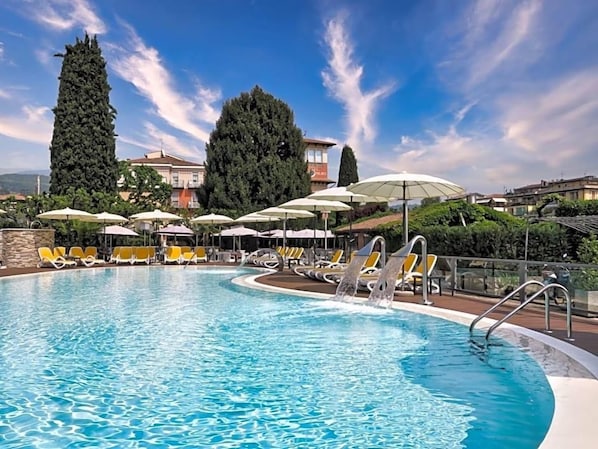 A swimming pool with sun loungers and umbrellas, surrounded by trees and buildings. - Villa Mulino (Garda)