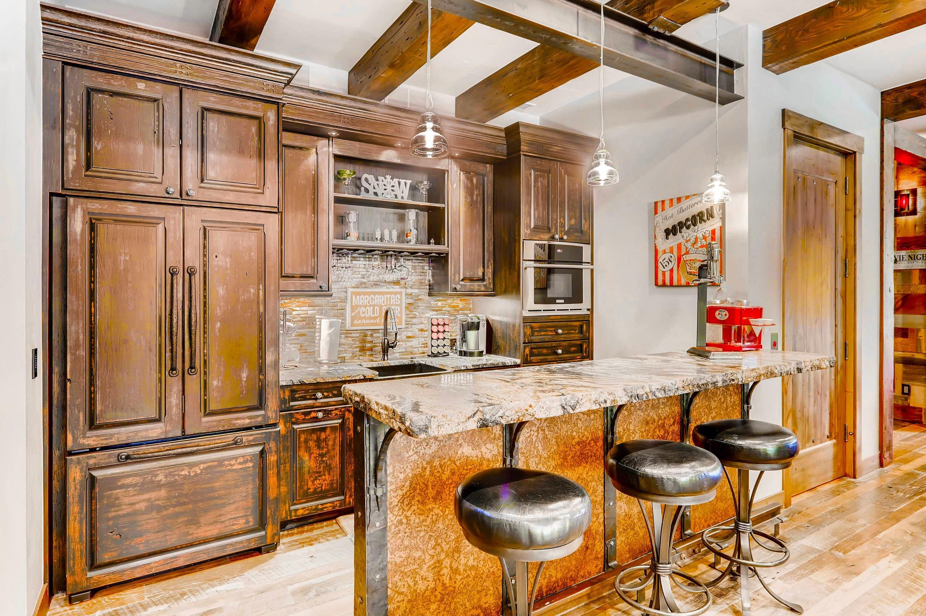 A kitchen with wooden cabinets, a marble island with bar stools, pendant lights, and a popcorn sign.