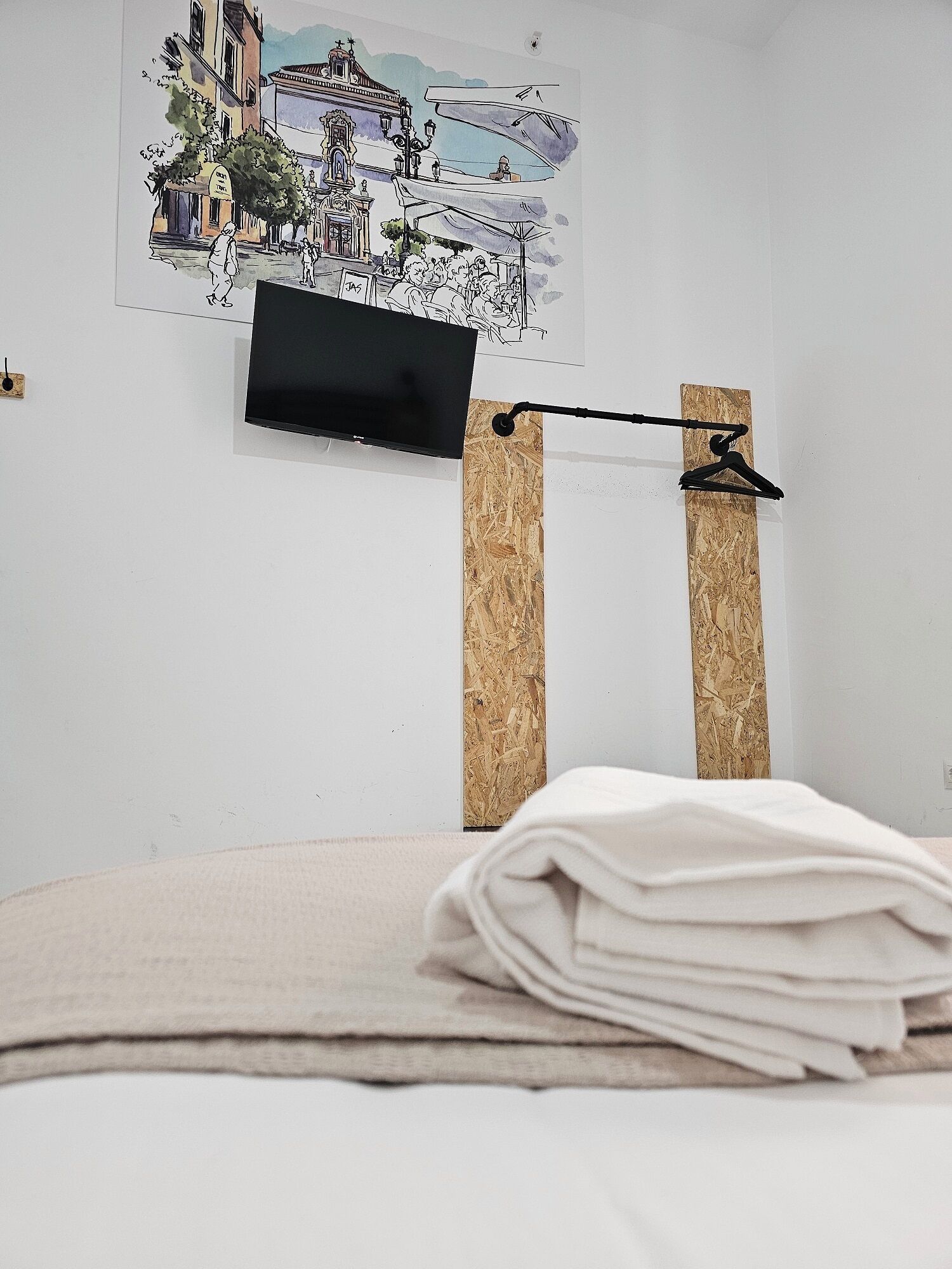 A hotel room with a bed, a television mounted on a wooden panel, and a wall-mounted artwork depicting a cityscape.