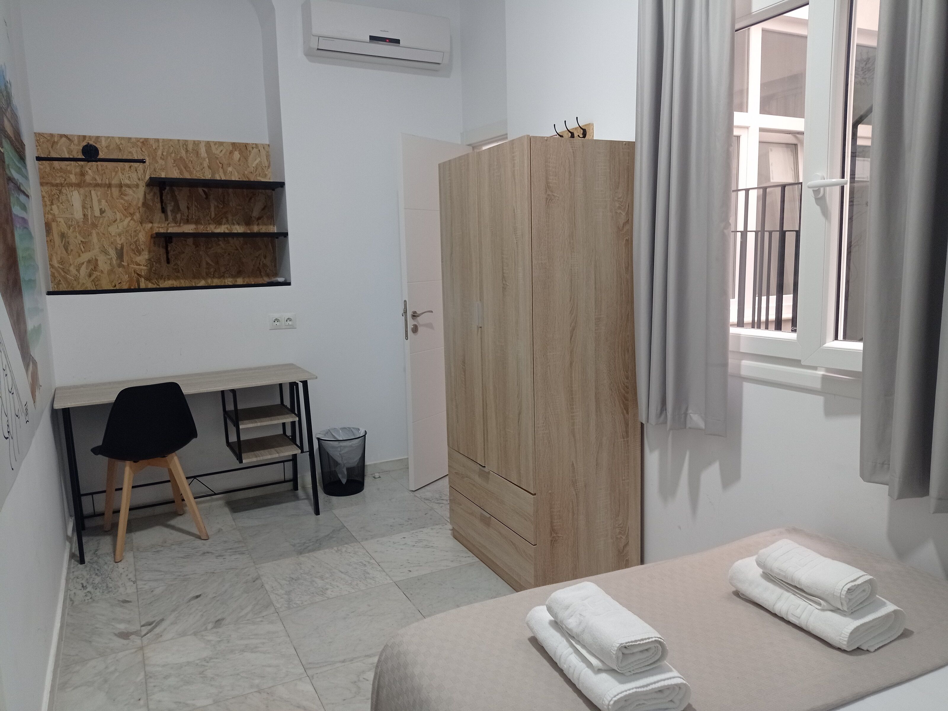 A bedroom with a bed, a wooden wardrobe, a desk with a chair, and a window with curtains.