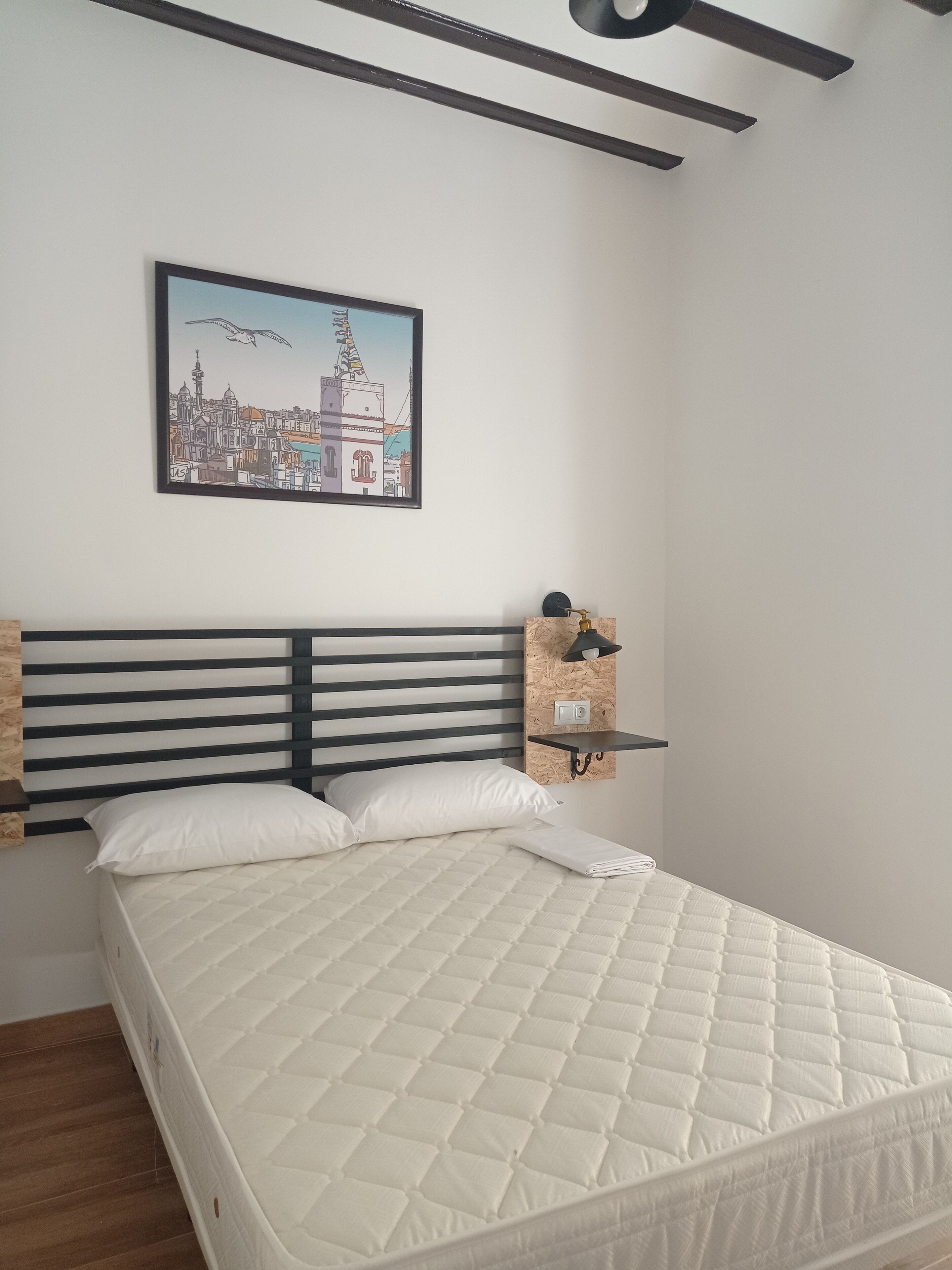 A neatly made bed with a quilted mattress, a headboard with horizontal slats, a wall-mounted shelf with a lamp, and a framed cityscape artwork above the bed.