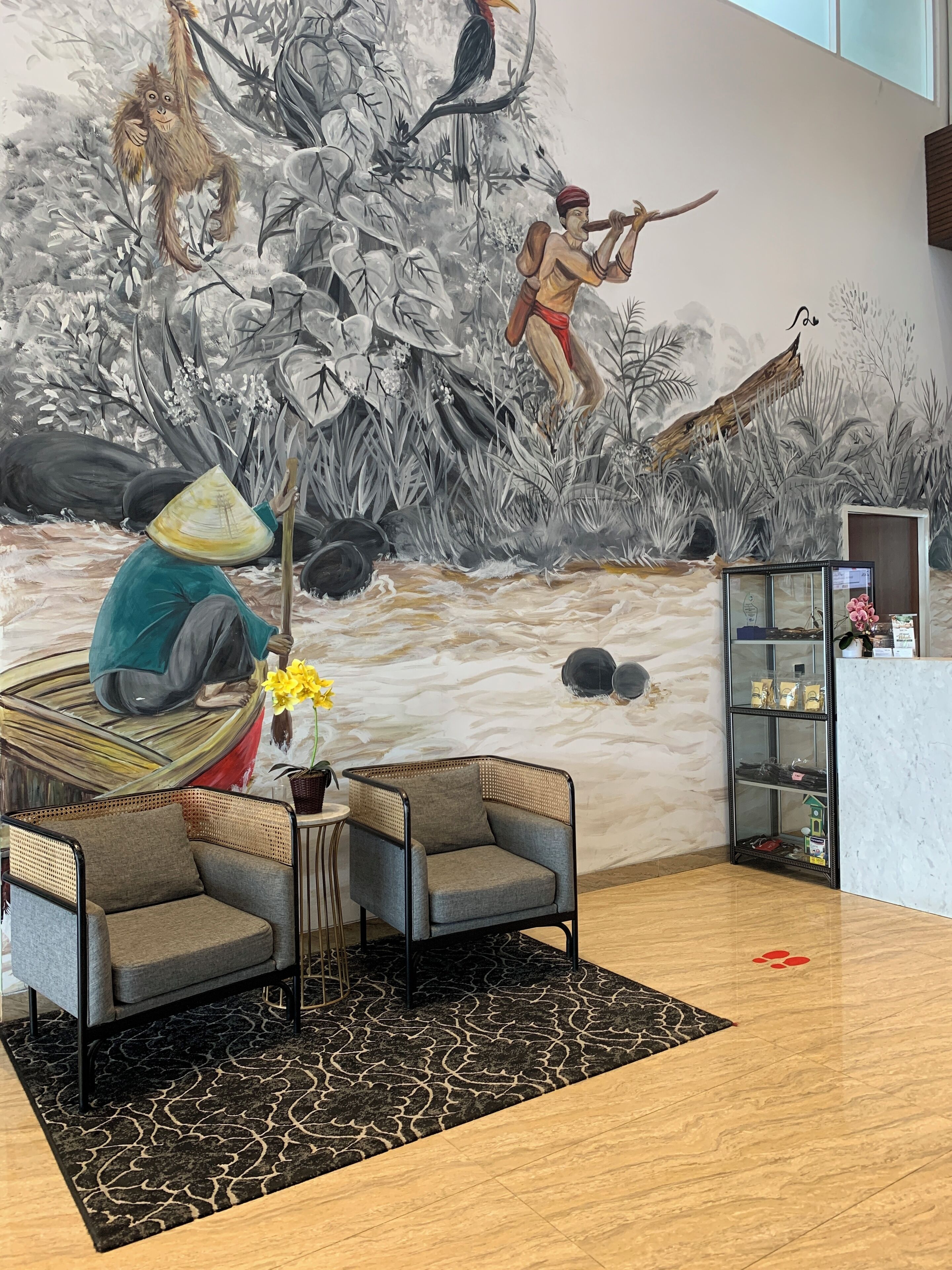 Quarto com um mural de uma cena de selva, dois poltronas cinzentas, uma pequena mesa com um vaso de flores e uma estante com diversos objetos.