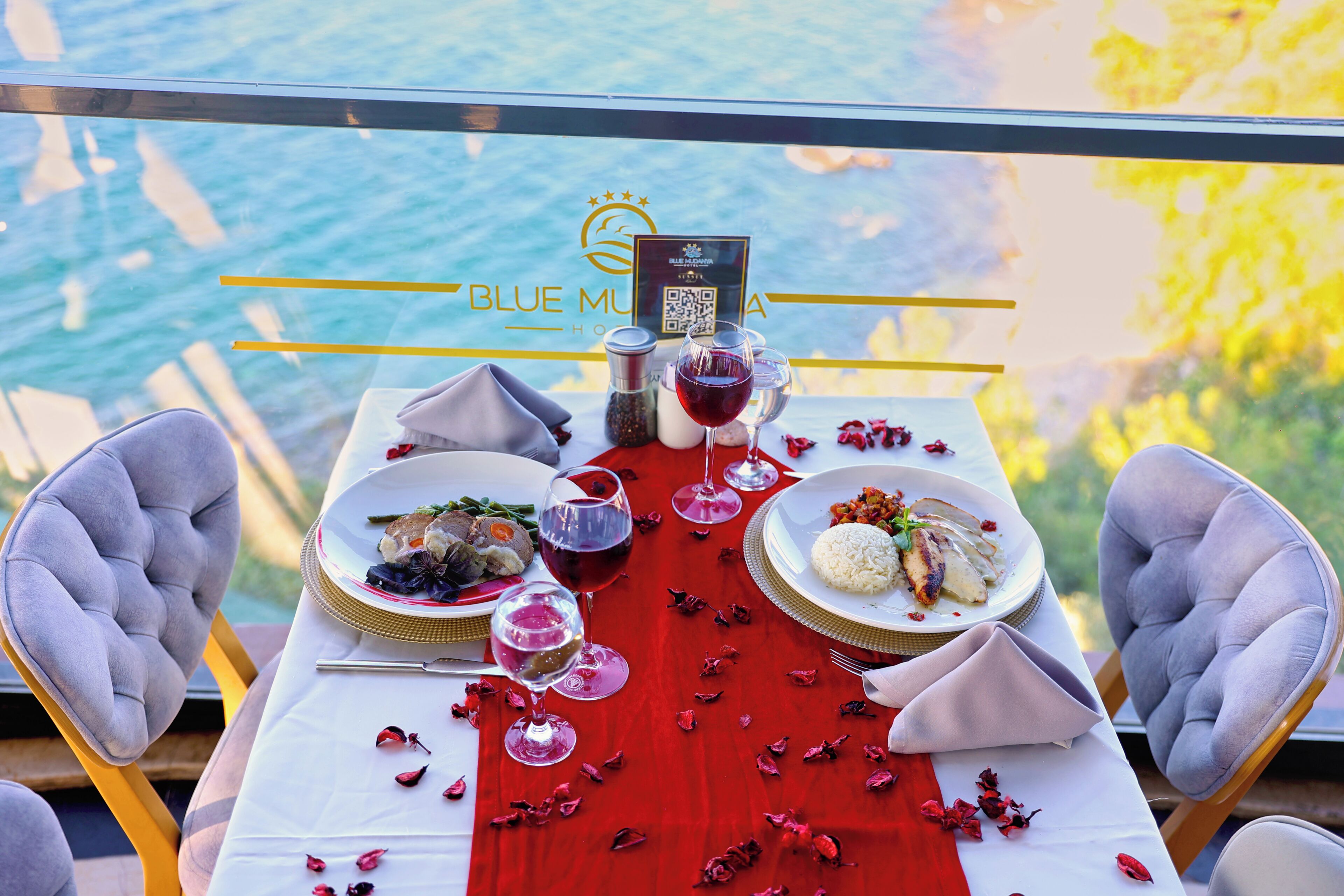 Une table dressée avec des verres à vin, des assiettes remplies de nourriture et un chemin de table rouge, offrant une vue sur la mer à travers une balustrade en verre.