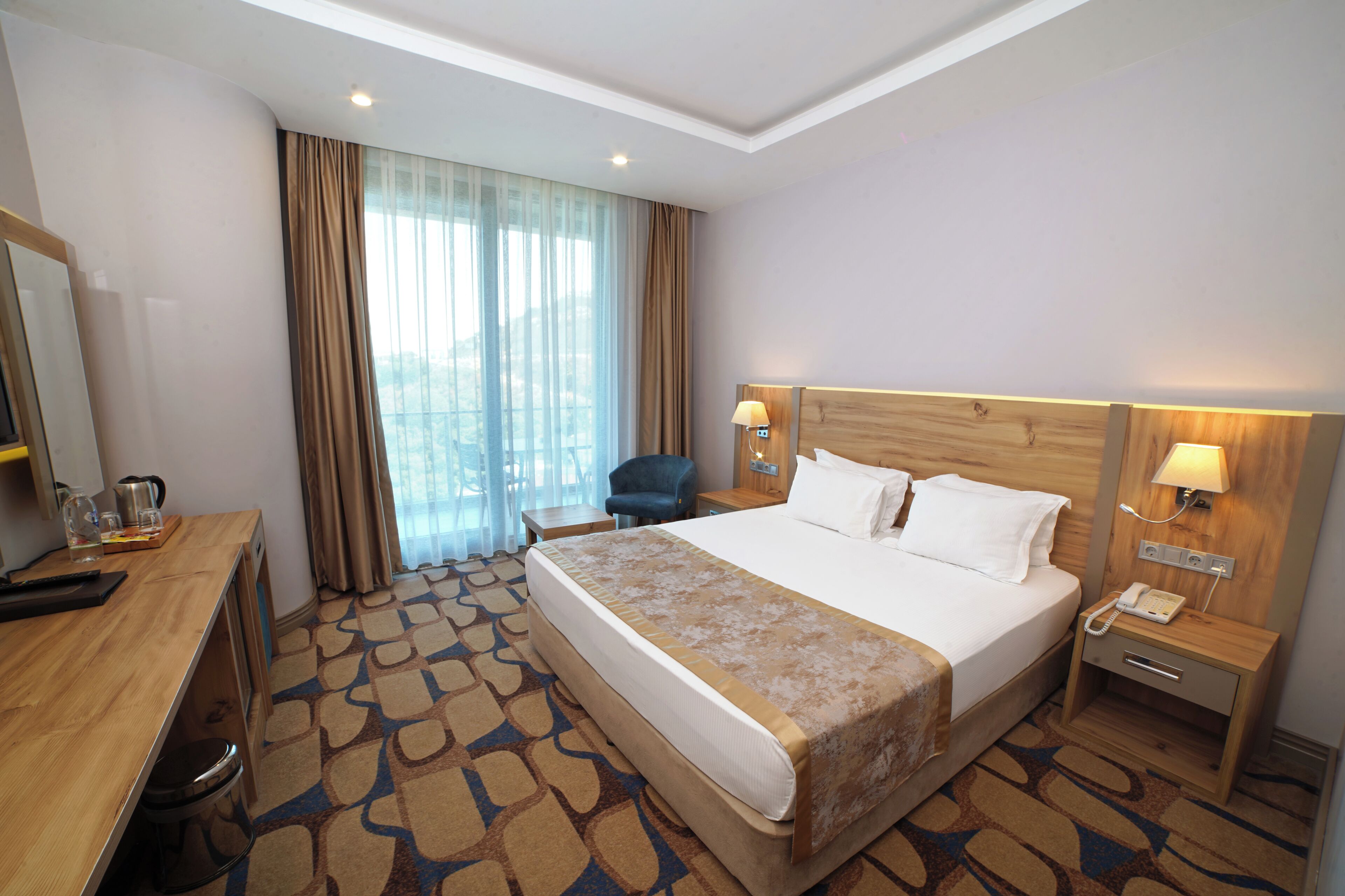 A hotel room with a bed, a desk, a chair, a nightstand with a phone, and a window with curtains.