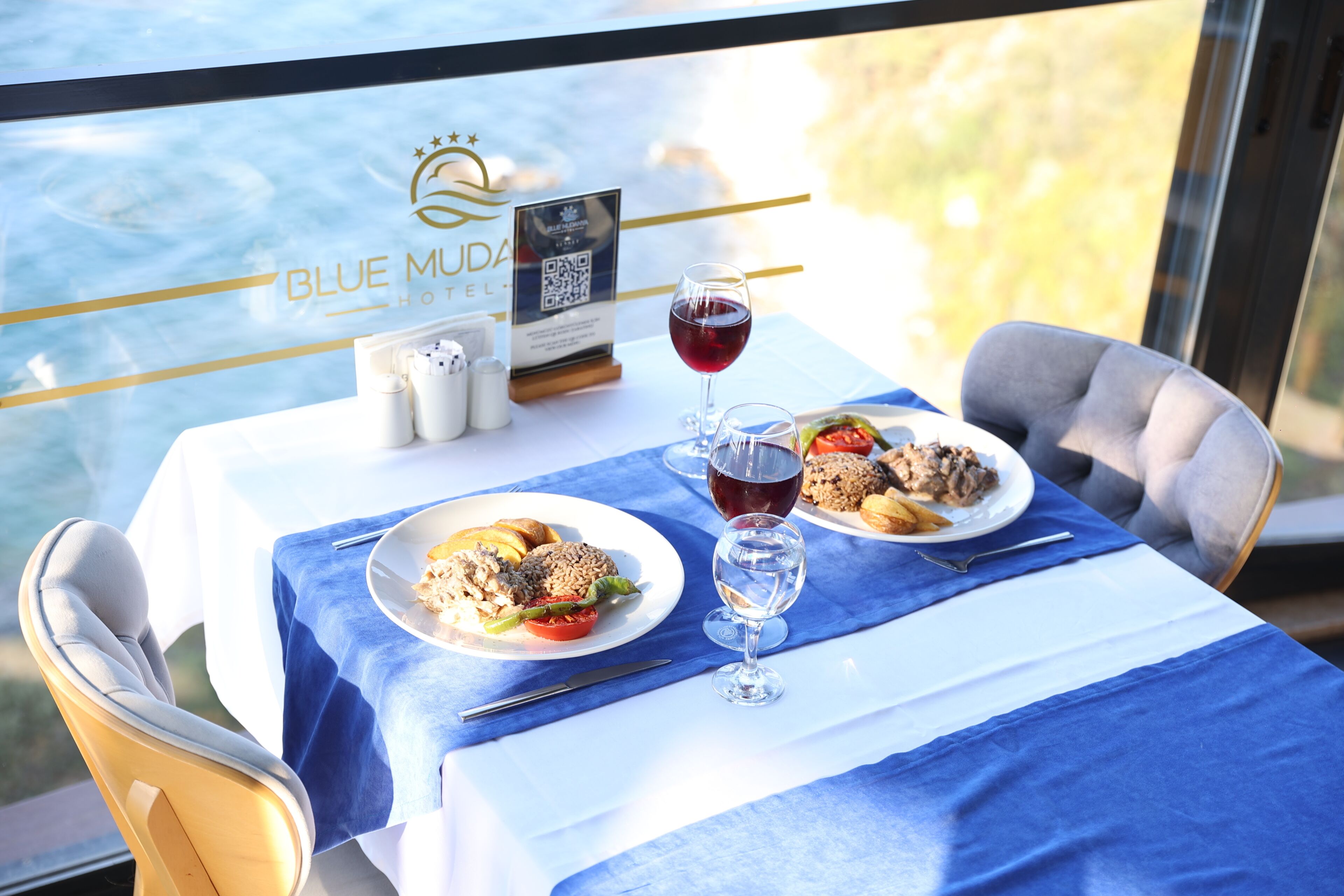 Una mesa de comedor con platos de comida, copas de vino tinto y un cartel con un código QR.