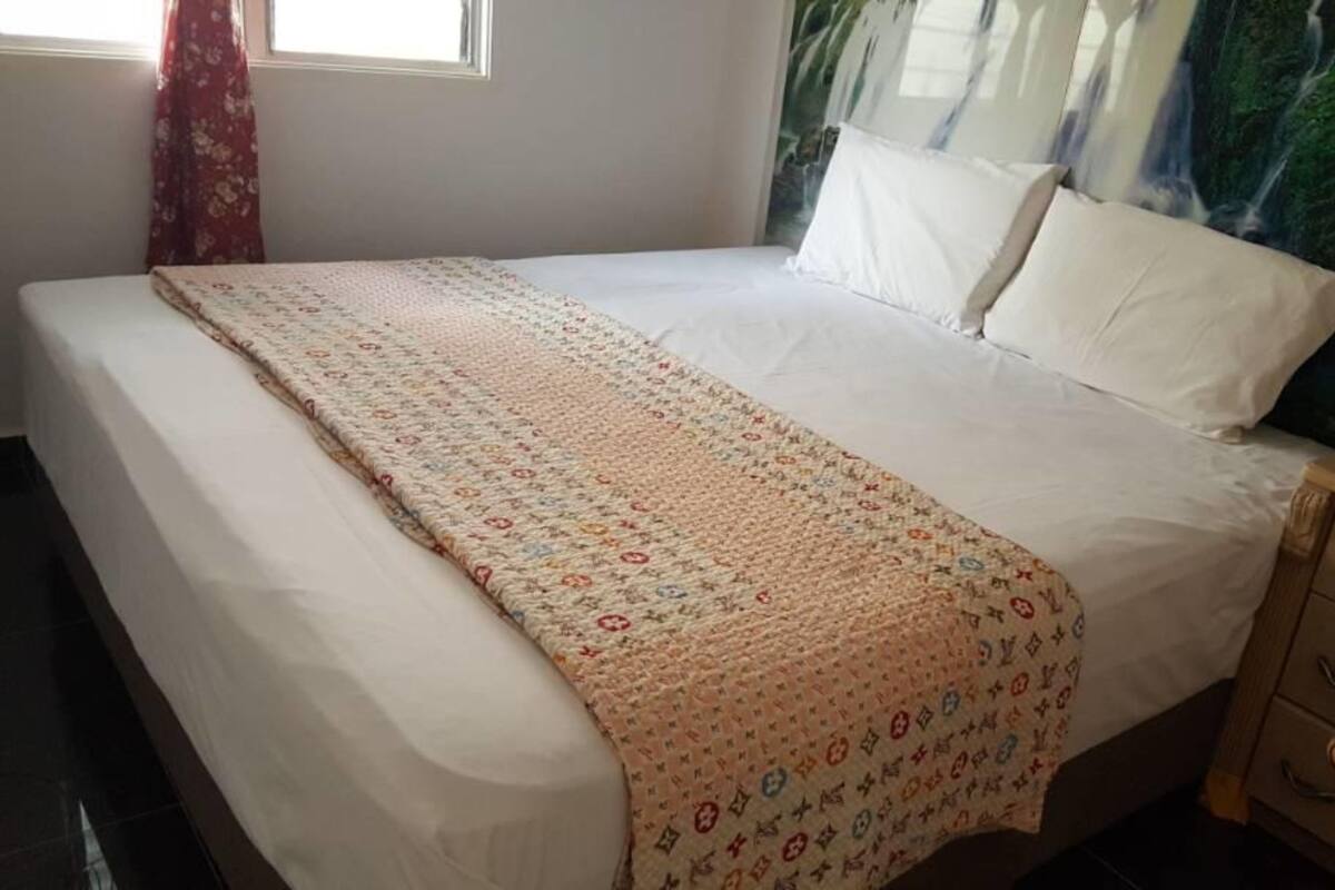 A neatly made bed with a patterned blanket, a wooden nightstand with drawers, and a wall with a nature-themed painting.
