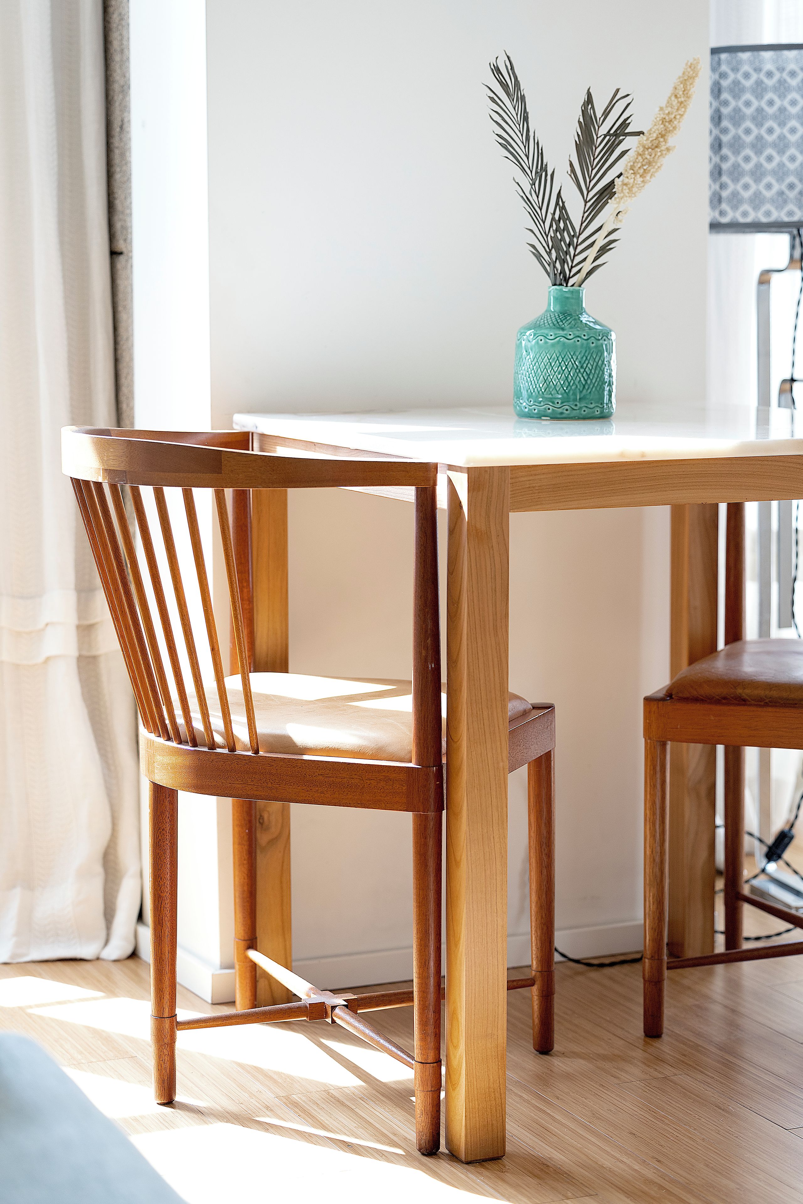 A wooden dining table with two chairs, a vase with dried plants, and a white wall in the background.