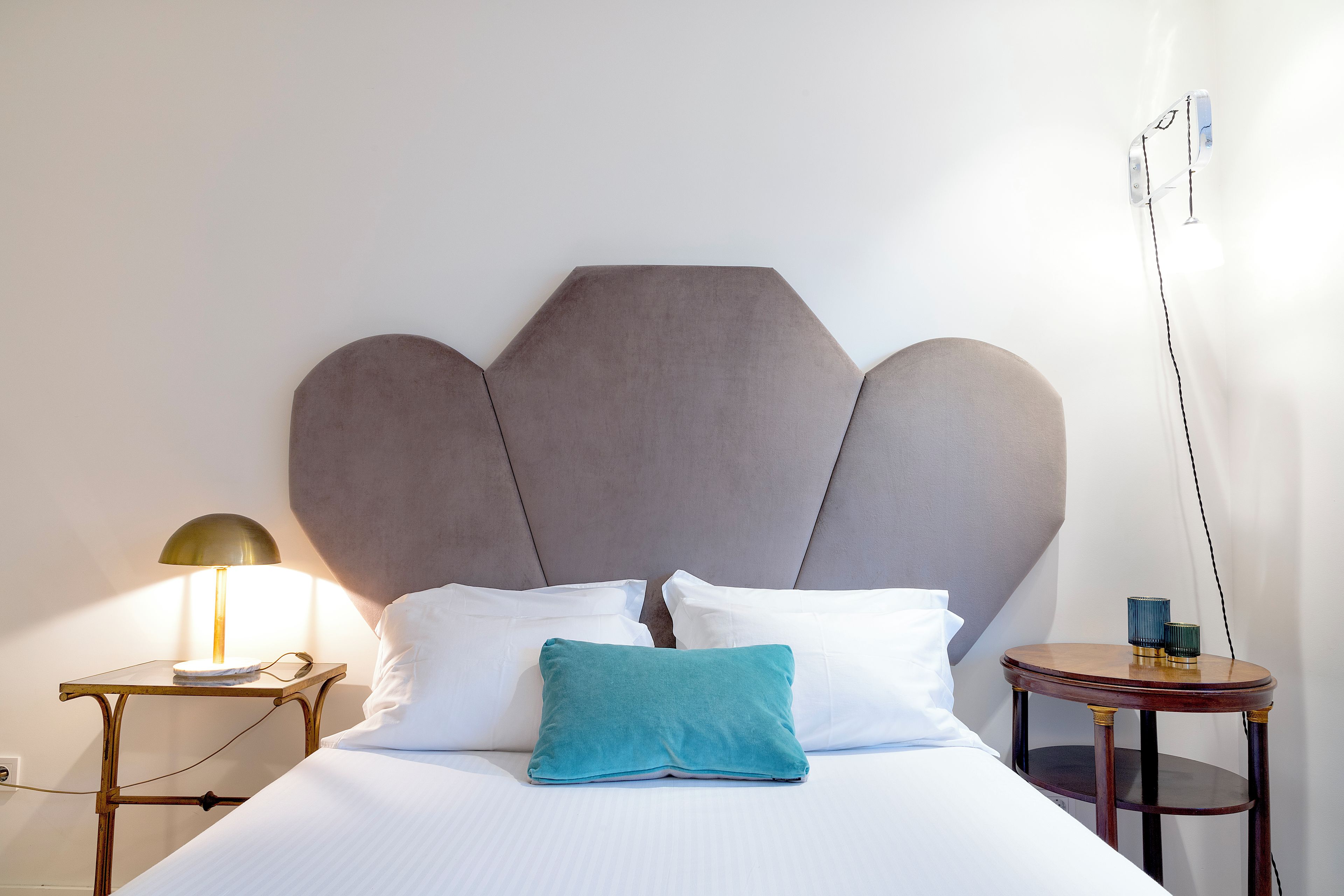 A neatly made bed with white linens and a teal pillow, a bedside lamp, and a small round table with two blue cups.