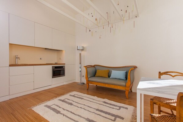 A modern kitchen with white cabinets, a wooden countertop, and a built-in oven. There is a wooden sofa with cushions, a white dining table with chairs, and a floor lamp. - The Art Porto (Porto)