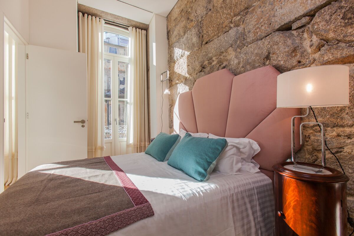 A bedroom with a stone wall, a bed with a pink headboard, teal pillows, and a bedside lamp.