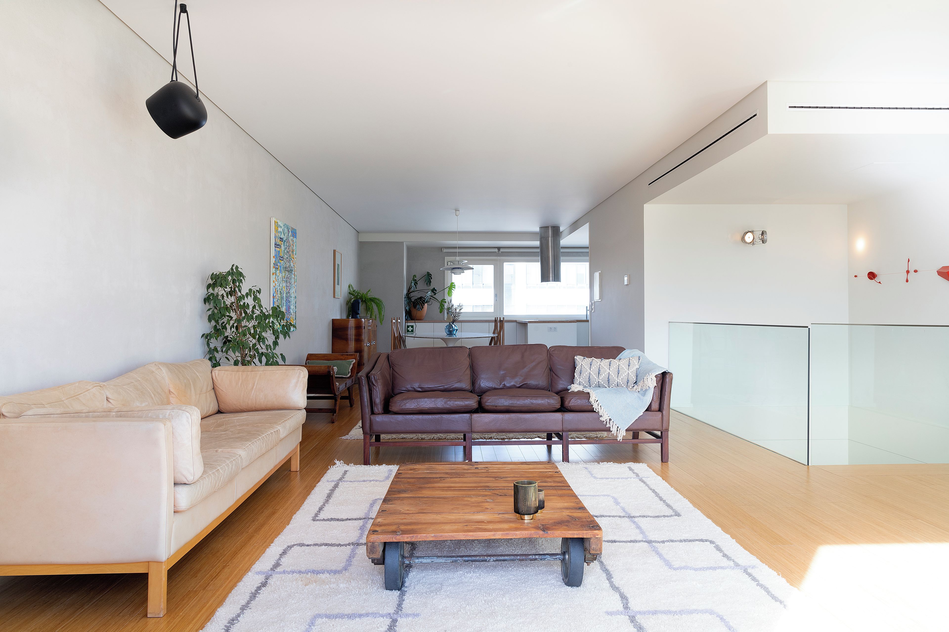 Ein modernes Wohnzimmer mit einem braunen Ledersofa, einem Couchtisch aus Holz und einem beigefarbenen Sofa. Im Hintergrund ist eine Küchenzeile mit einem Esstisch und Stühlen zu sehen.