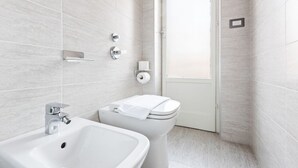 A modern bathroom with a white sink, toilet, and shower area, featuring a tiled wall and a door leading to another room. - GuestHost - Lovely studio apartment complete with all major amenities (Milano)
